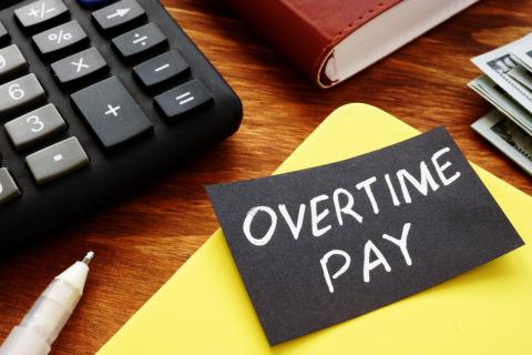 A photograph of a calculator on a desk. There's a yellow folder next to it with a sticky note on top that says "overtime pay."