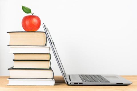 An open laptop with the screen resting against a stack of six books, which has an apple perched on top.