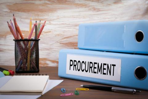 A photograph of two binders stacked on a table. One binder has the word "procurement" written on it. There is a pen, four paper clips, a notepad resting on sheets of paper, two highlighters, and a cup of pencils next to the binders.