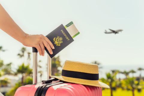 A person holding a U.S. passport that has a flight boarding pass tucked inside. The person is resting their hand on the handle of a suitcase, which has a hat perched on top of it. An airplane is taking off in the background.