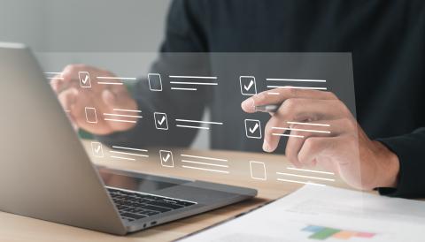 A photograph of a laptop with a virtual checklist screen in front of it. The computer user is holding a pen and marking off check boxes.