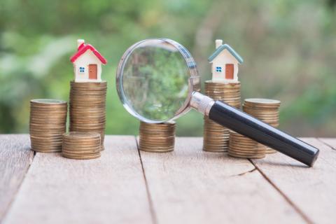 A photograph of a magnifying glass over two small houses sitting on stacks of coins.